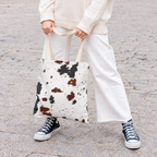Woman holding the black and brown cow print tote bag in front of her on a street