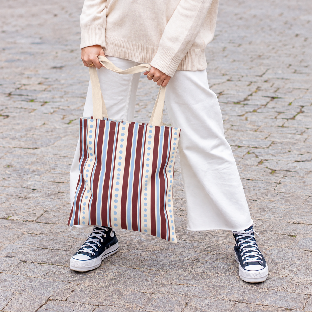 Woman in white trousers holding the blue burgundy dot striped tote bag by its handles