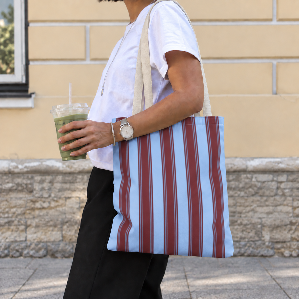Woman carrying the blue burgundy striped tote bag on her shoulder while holding an iced drink