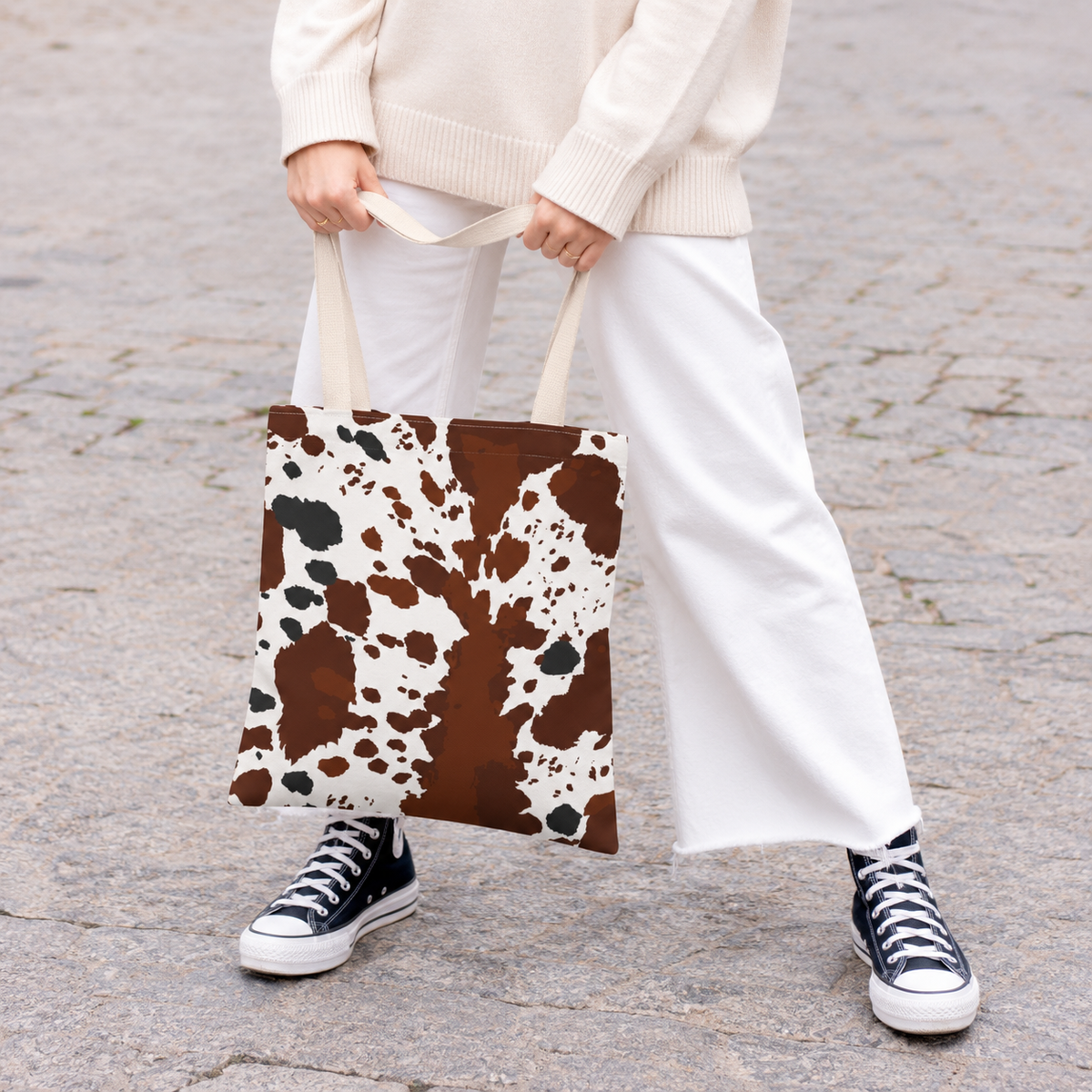 Woman holding a brown white cow print tote bag with white trousers and sneakers