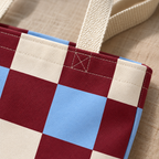 Close-up of the burgundy, light blue and cream checks and the beige handle on the checkered tote bag