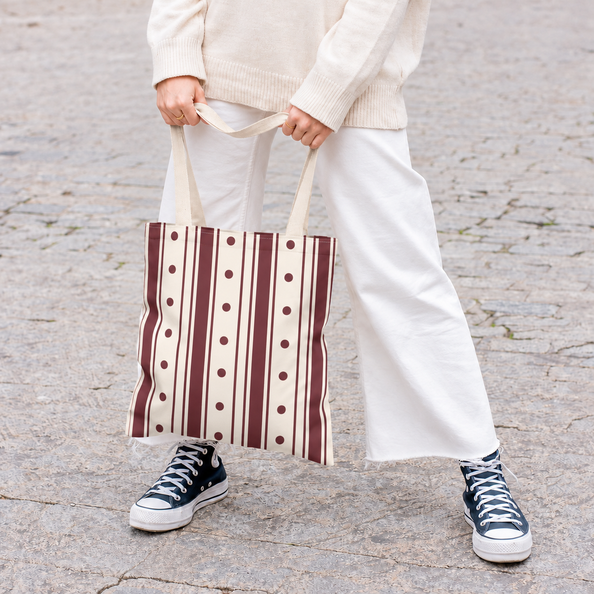 Woman in white trousers holding the burgundy polka dot striped tote bag by its handles