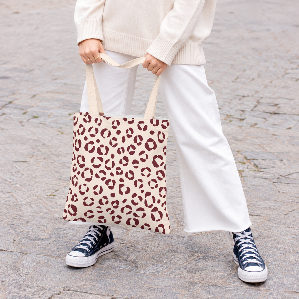 Woman holding a cream tote bag with burgundy leopard spots, white trousers and sneakers