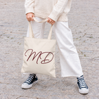 Model in white trousers holding the cream personalized tote bag with burgundy script initials