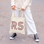 Woman in white trousers and sneakers holding the cream RS personalized tote bag