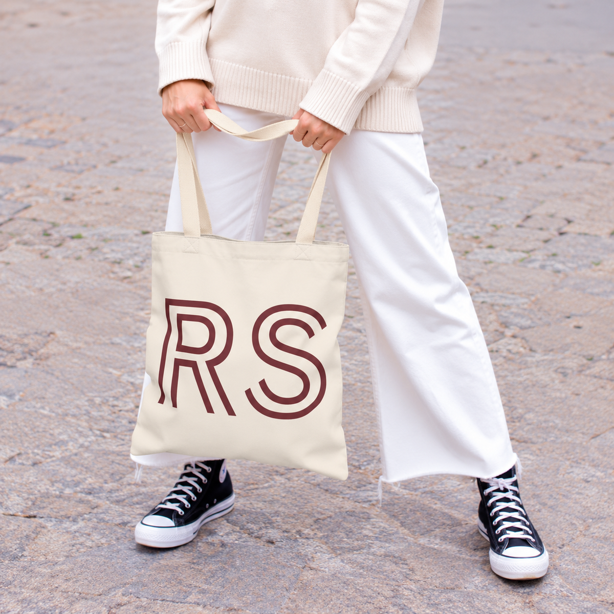 Woman in white trousers and sneakers holding the cream RS personalized tote bag