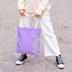 Model holding the Lavender Green Name personalized tote bag in front of white trousers
