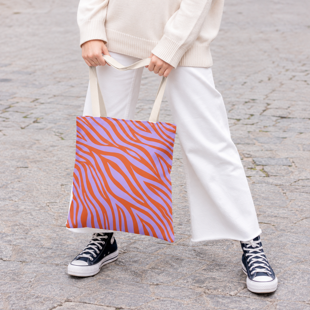 Woman in white trousers and sneakers holding the lavender orange zebra print tote bag