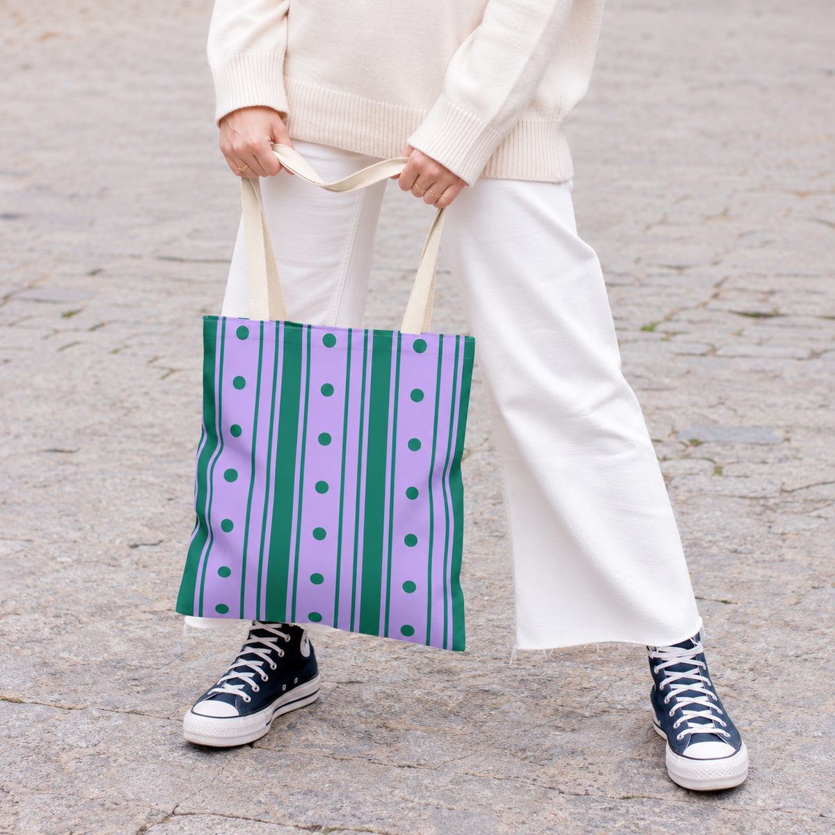 Woman in white trousers and sneakers holding the purple green striped tote bag