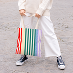 Woman in white trousers and sneakers holding the red green lavender striped tote bag