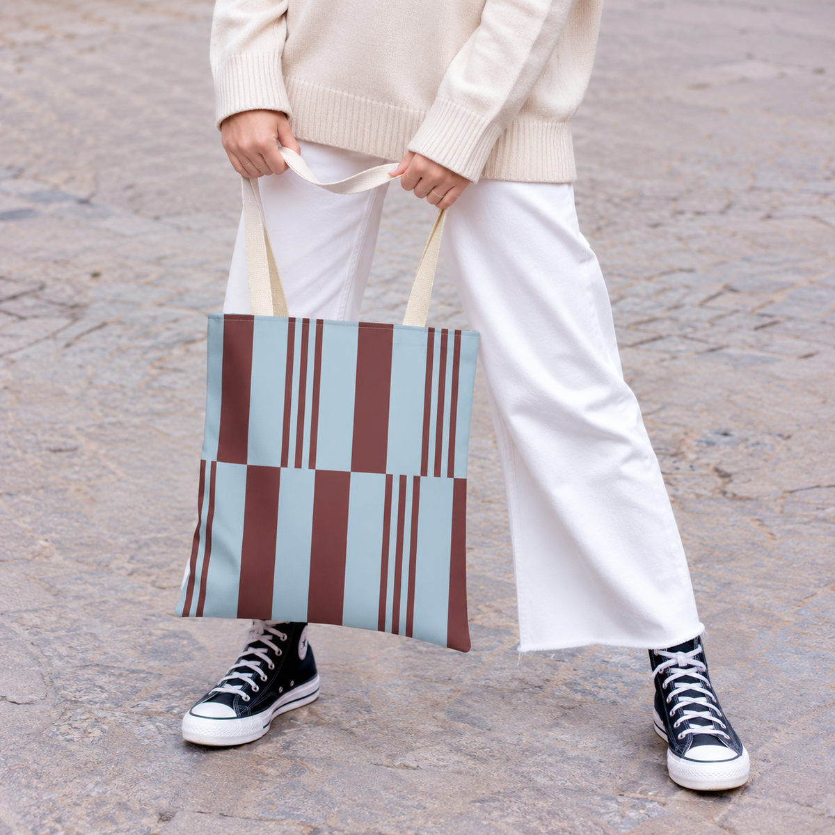 Model holding the sky blue burgundy block striped tote bag in front of white trousers